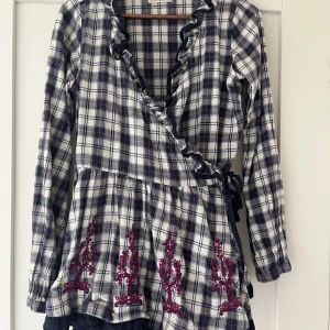 Rutig omlottblus/tunika Odd Molly - Supersöt omlottblus från Odd Molly med rutig design i vitt, marinblått och rosa. Långärmad med volangkant och knyt i sidan. Broderade detaljer i vinrött nedtill ger extra vibe. Perfekt för dig som gillar unika detaljer och skön bomullskänsla.