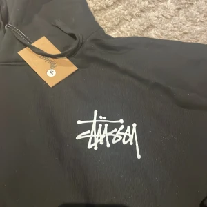 Svart Stüssy hoodie - Svart hoodie från Stüssy i storlek S med stor vit graffiti-inspirerad logga på både bröstet och ryggen. Tröjan har huva med snörning och ribbade muddar vid ärmslut och nederkant. Skulle säga att de är en storklek M