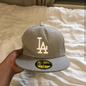 Grå LA Dodgers keps - Snygg grå LA Dodgers 59FIFTY keps från New Era med vit broderad LA-logga framtill och World Series 1988-detalj på sidan. Klassisk rak skärm och MLB-logga baktill. Perfekt för dig som gillar streetwear och sportig stil. Endast provad säljer för att den är för står.