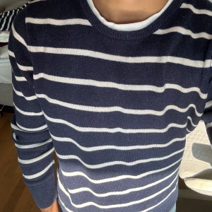 Randig marinblå stickad tröja - Säljer en stilren marinblå stickad tröja med vita horisontella ränder. Tröjan har rund halsringning och långa ärmar med ribbade muddar. Perfekt för dig som gillar en klassisk och avslappnad look.
