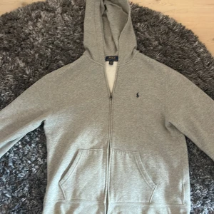 Grå hoodie från Polo Ralph Lauren  - Säljer en grå hoodie från Polo Ralph Lauren i storlek XL (barnstorlek). Skulle säga att den motsvarar en XS i vanlig storlek. Tröjan har huva, dragkedja framtill och två stora fickor. Klassisk Polo-logga broderad på bröstet. Perfekt för chill dagar och har en mjuk insida i bomullsmix.  Finns en liten fläck på baksida ryggAnvänd mycket men ändå bra skick.