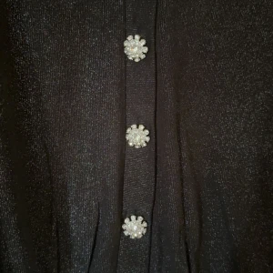 Glittrig svart kofta med pärlknappar - Supersnygg svart glittrig kofta från Sisters Point med v-ringning och långa ärmar. Koftan har fem blommiga pärlknappar som ger extra bling och festkänsla. Materialet är mjukt och skimrande, perfekt för att sticka ut. Passar dig som gillar att glänsa lite extra.