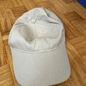 Beige keps med justerbar rem - Snygg beige keps med klassisk böjd skärm och ventilerande hål upptill. Kepsen har en enkel design utan logga och är tillverkad i bomullstyg. Justerbar rem baktill för perfekt passform. Perfekt för dig som gillar stilrena accessoarer.