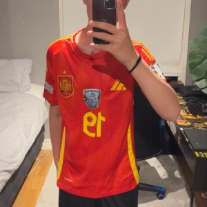 Spansk Lamine Yamal tröja - Säljer en röd Spanien fotbollströja från Adidas i slim fit, storlek S. Tröjan har gula detaljer, nummer 19 på framsidan och Nations League-vinnarmärke. tillverkad i lätt polyester med ventilerande paneler. Svinsnygg Lamine Yamal fotbollströja och ett väldigt bra skick💯