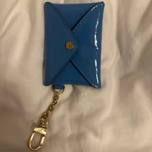 Blå mini plånbok i lack med guldkedja - Snygg blå plånbok i glansigt lackmaterial med kuvertformad design och tryckknapp. Utrustad med guldfärgad kedja och karbinhake, perfekt att fästa på väskan eller nyckelknippan. Passar bra för mynt eller småsaker när du vill ha något stilrent och annorlunda