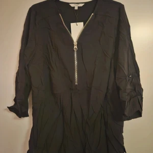 Svart topp med dragkedja Cellbes - Svart topp från Cellbes med snygg silverfärgad dragkedja framtill och v-ringad hals. Modellen är långärmad och har en loose passform. Perfekt för dig som gillar enkel stil med coola detaljer. Materialet känns lätt och luftigt.