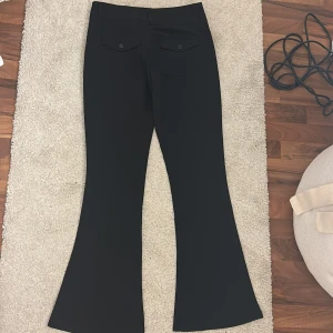 Svarta bootcut-byxor från Gina Tricot - Svarta bootcut-byxor från Gina Tricot med snygg passform och vida ben. Byxorna har två bakfickor med lock och knappar, bälteshällor och stängs med dragkedja och knappar framtill. Perfekta för en clean och stilren look.
