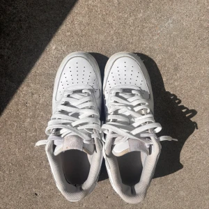 Nike Air Force 1 vita sneakers - Klassiska Nike Air Force 1 sneakers i helvitt skinn med perforerad tå, rund tå och platt sula. Swoosh-logga på sidan och snörning framtill. Perfekta för dig som gillar en clean och tidlös look.