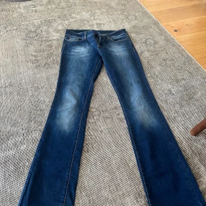 Blå bootcut jeans med knappar - Snygga blå jeans med bootcut-modell och slitningar på låren. Jeansen har klassiska fem fickor, synliga knappar i gylfen och kontrastsömmar. Perfekta för dig som gillar en retrovibe och vill ha ett par jeans med lite extra attityd.
