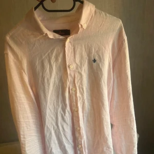 Ljusrosa linneskjorta från Morris - Snygg ljusrosa skjorta från Morris i linne med klassisk krage och knappar framtill. Skjortan har lång ärm och en liten blå broderad logga på bröstet. Perfekt för en stilren och avslappnad look.