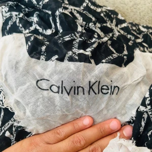 Svartvit Calvin Klein halsduk/ sjal - Snygg halsduk/ sjal från Calvin Klein i svart och vitt med grafiskt CK-mönster över hela tyget. Lätt och luftig känsla, perfekt att slänga över axlarna. Fransiga kanter och stor logga i ena hörnet ger en cool touch.
