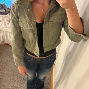 Olivgrön croppad jeansjacka S - Snygg croppad jeansjacka i olivgrönt från Gina tricot, Perfect Jeans. Jackan har klassisk krage, två bröstfickor med lock och silverfärgade knappar framtill. Perfekt för dig som gillar en avslappnad men trendig stil.