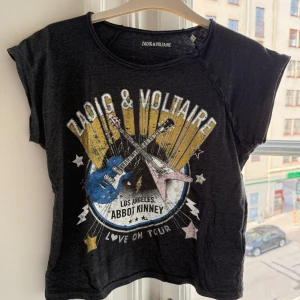 Mörkgrå t-shirt Zadig & Voltaire - Cool mörkgrå t-shirt från Zadig & Voltaire med färgstarkt tryck av två elgitarrer och texten 'Love On Tour'. Rund hals, korta ärmar och dekorativa knappar längs axeln. Materialet är mjukt med 60% bomull och 40% polyester. Perfekt för dig som gillar rockig stil.