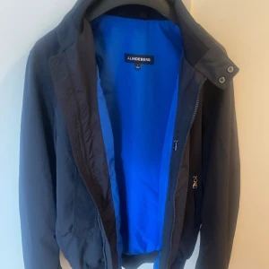 Svart vindjacka från J.Lindeberg - Svart vindjacka från J.Lindeberg i storlek S. Jackan har en stilren design med hög krage, dragkedja och tryckknappar framtill. Fodret är blått för en snygg kontrast. Perfekt för dig som gillar minimalistisk och sportig stil. Den är använd fåtal gånger och ser ny ut!  