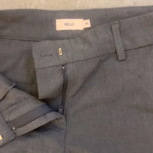 Grå kostymbyxor från Nelly - Stilrena grå kostymbyxor från Nelly med raka ben och klassiska fickor. Byxorna har hällor för bälte och stängs med dragkedja och hake. Perfekta för en clean och dressad look.