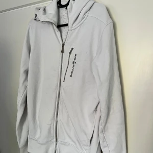 Vit hoodie från Sail Racing med dragkedja - Säljer en vit hoodie från Sail Racing i storlek S. Tröjan har hel dragkedja, huva och en snygg bröstficka med logotyp. Mjuk insida och klassisk passform, perfekt för chill eller häng. Grym att matcha med jeans eller joggers.