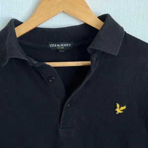 Svart pikétröja från Lyle & Scott - Svart pikétröja från Lyle & Scott med klassisk krage och två knappar framtill. Tröjan har korta ärmar och den ikoniska gula fågeln broderad på bröstet. Perfekt för dig som gillar stilrena och sportiga plagg med snygga detaljer.