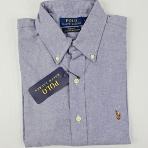 Blå skjorta från Polo Ralph Lauren - Klassisk blå skjorta från Polo Ralph Lauren i slim fit med button-down krage och broderad logga på bröstet. Skjortan är tillverkad i 100% bomull och har vita knappar framtill. Perfekt för en stilren och avslappnad look.