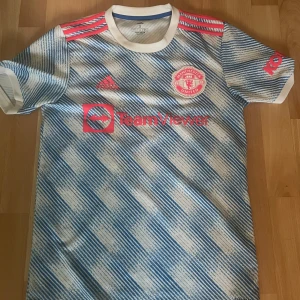 Äkta Manchester United bortatröja Adidas S - Manchester United bortatröja från Adidas i storlek S. Tröjan har ett randigt mönster i vitt och blått med röda detaljer och klubbmärke på bröstet. Tillverkad i Primegreen-material med klassisk rund hals och korta ärmar. Snygga röda Adidas-ränder på axlarna. Den är helt äkta köpt på unisport