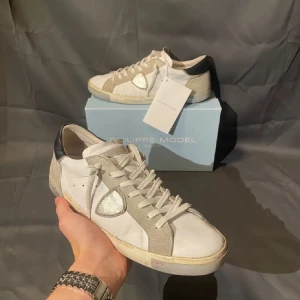 Philippe Model vita sneakers med grå detaljer - Snygga vita sneakers från Philippe Model med grå mockadetaljer och svart häl. Skorna har en ikonisk sköld i silver på sidan, vita snören och platt sula. Perfekta för dig som gillar stilrena men ändå unika sneakers med exklusiv känsla.