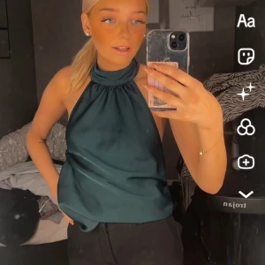 Mörkgrön halterneck linnetopp - Snygg mörkgrön halterneck topp i glansigt material med hög hals och ärmlös design. Toppens tyg faller mjukt och har en elegant, enkel look som passar perfekt till jeans eller kjol. Stängs med dragkedja i sidan för en smidig passform. Ena knappen har lossnat för att knäppa i nacken. 
