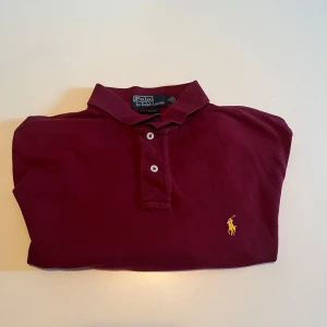 Vinröd Polo Ralph Lauren Piké - Snygg vinröd t-shirt från Polo Ralph Lauren i klassisk pikémodell med krage och två knappar.  Tröjan är i storlek XL men passar L också. Varan är äkta såklart.  Kan mötas i Stockholm eller frakta 🚚 