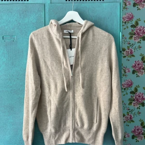 Beige hoodie i kashmir från Soft Goat - Snygg beige hoodie från Soft Goat i mjuk kashmir. Tröjan har dragkedja framtill, huva med snörning. Förgen är Natural sand. Helt ny, med prislapp. Köpt för 2500kr.