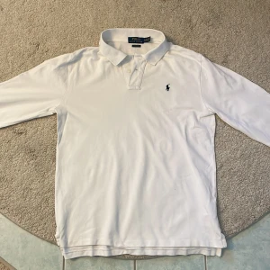 Vit långärmad pikétröja Ralph Lauren - Vit långärmad pikétröja från Polo Ralph Lauren i klassisk passform. Tröjan har krage, två knappar vid halsen och den ikoniska broderade loggan på bröstet. Tillverkad i mjuk bomull som är skön mot huden. Perfekt för dig som gillar clean och stilren look.