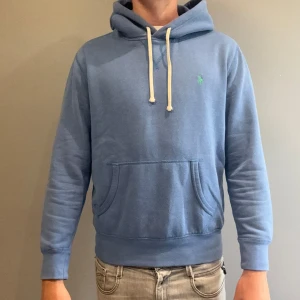 Blå Ralph Lauren hoodie - Snygg blå hoodie från Polo Ralph Lauren med klassisk grön logga på bröstet. Tröjan har huva med vita snören, stor magficka och ribbade muddar vid ärmar och nederkant. Perfekt för chill dagar och enkel att matcha med jeans eller joggers. Han på bild är 185, 74kg!