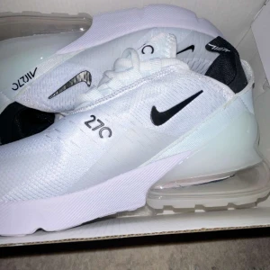 Nike Air Max 270 vit/svart strl 37.5 - Nike Air Max 270 sneakers i fräsch vit färg med svarta detaljer och stor synlig Air-enhet i hälen. Ovandel i mesh för bra andningsförmåga, klassisk snörning och modern design. Perfekta för dig som gillar streetstyle och bekväma skor.