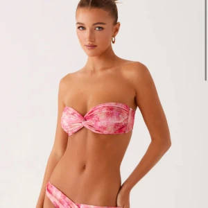 Rosa bandeau bikini med twist - Säljer en snygg bandeau bikini i rosa och ljusrosa toner med twistad detalj framtill. Bikinin har ett abstrakt mönster och axelbandslös överdel. Materialet är mjukt och stretchigt, perfekt för strandhäng och soliga dagar.