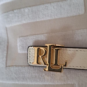 Vändbart Vit / pink skinnbälte från Ralph Lauren - Snyggt vitt och pink färgat vändbart  bälte från Ralph Lauren i präglat skinn med guldfärgat RL-spänne och detaljer. Smal 2 cm och stilren design som ger en lyxig touch till din outfit. Perfekt för dig som gillar klassiska accessoarer med exklusiv känsla. Totallängd 90cm. Innersta hålet på 70cm yttersta 80cm med 3 stycken mellanliggande hål. Äkta skinn.
