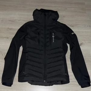 Svart Sail Racing jacka  - Originalpris är 3500kr! Säljer en svart pufferjacka från Sail Racing med huva och dragkedja framtill. Jackan har två bröstfickor med dragkedja, quiltad front och rygg samt släta ärmar. Perfekt för kyliga dagar och snygg streetstil.