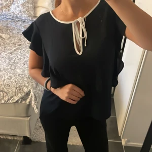 Svart blus med volang och knyt - Säljer en svart blus med vita detaljer vid halsringningen och knyt framtill. Blusen har korta volangärmar som ger en snygg och luftig känsla. Perfekt för dig som gillar stilrena plagg med en twist.