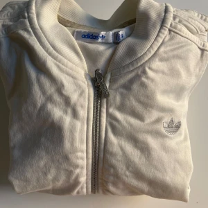 Vit bomberjacka från Adidas med stenar - Snygg vit bomberjacka från Adidas med dragkedja framtill och ikoniska loggan i silver på bröstet. Baksidan har stora Adidas-loggan dekorerad med glittriga stenar för extra bling. Jackan har ribbade muddar och är långärmad, perfekt för en sportig och trendig look.