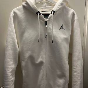 Vit hoodie från Jordan med dragkedja - Säljer en fräsch vit hoodie från Jordan med svart Jumpman-logga på bröstet och texten '23ENGINEERED' på ryggen. Hoodien har snörning i huvan, hel dragkedja och långa ärmar. Perfekt för chill eller streetwear. Materialet är mjuk bomull och polyester.