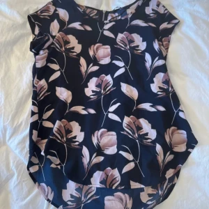 Blommig blus i mörkblått och rosa - Supersnygg blus med mörkblå botten och stora blommor i ljusrosa och beige toner. Modellen är ärmlös med rund halsringning och dragkedja bak. Perfekt för dig som gillar ett romantiskt och trendigt uttryck.