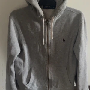 Grå hoodie från Polo Ralph Lauren - Snygg grå hoodie från Polo Ralph Lauren med dragkedja och huva med vita snören. Tröjan har lång ärm, två fickor fram och klassisk Polo-logga broderad på bröstet. Perfekt för chill dagar och enkel att matcha med jeans eller joggers.