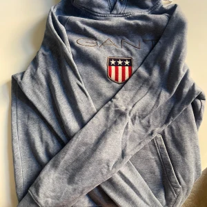 Blå hoodie från GANT med broderad logga - Säljer en snygg ljusblå hoodie från GANT med broderad logga och sköld på bröstet. Tröjan har huva med snörning och en klassisk känguruficka framtill. Perfekt för chill dagar och enkel att matcha med jeans eller joggers.