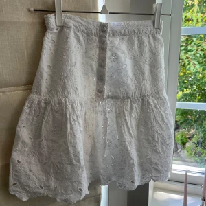 Vit broderad kjol Holly & Whyte XS - Supersöt vit kortkjol från Holly & Whyte med broderade blommor och knappar framtill. Kjolen har volangkant och resår i midjan för skön passform. Perfekt för sommarens alla dagar, lätt och luftig i bomull.