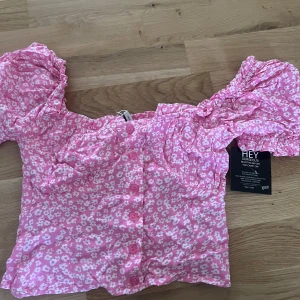 Rosa blommig blus från NLY Trend - Supersöt rosa blus med vita blommor från NLY Trend. Modellen har puffiga korta ärmar, knappar framtill och en croppad passform. Perfekt för dig som vill ha en girly och trendig look. Materialet är mjukt och lätt bomull.