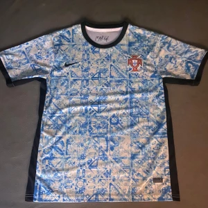 Portugal Nike fotbollströja blåmönstrad - Säljer en Portugal fotbollströja från Nike med unikt blått grafiskt mönster på vit bas. Tröjan har korta ärmar, svart krage och svarta detaljer längs sidorna. Portugal-emblemet sitter på bröstet och materialet är lätt och andas. Passar folk med size M-L