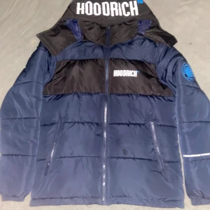 Blå pufferjacka från Hoodrich - Snygg mörkblå pufferjacka från Hoodrich med svart panel upptill och stora loggor både fram, bak och på huvan. Jackan har dragkedja, huva och coolt tryck på ärmen. Perfekt för dig som vill sticka ut med streetstyle och hålla dig varm. 