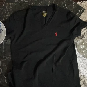  Äkta Svart v-ringad t-shirt Polo Ralph Lauren - Svart v-ringad t-shirt från Polo Ralph Lauren i custom slim fit. Klassisk röd broderad logga på bröstet. T-shirten är kortärmad och gjord i mjuk bomull, perfekt för en clean och stilren look.