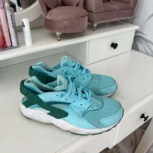Nike Air Huarache blå sneakers - Säljer ett par fräscha Nike Air Huarache sneakers i ljusblå och turkos färg med gröna detaljer. Skorna har mesh och syntetmaterial på ovansidan, snörning och en vit platt sula. Perfekt för dig som gillar sportig och cool stil.