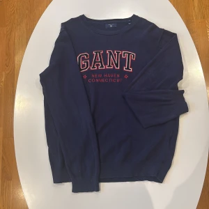 Marinblå stickad tröja från GANT XL - Snygg marinblå stickad tröja från GANT i storlek XL. Tröjan har broderad logga och texten 'New Haven Connecticut' i rött och vitt framtill. Rund hals och långa ärmar med ribbade muddar. Perfekt för dig som gillar klassisk college-stil.