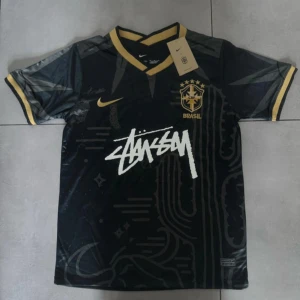 Svart Brasil x Stüssy x Nike fotbollströja - Unik svart Brasil-fotbollströja i samarbete med Stüssy och Nike. Tröjan har korta ärmar, guldfärgade detaljer vid krage och ärmslut, samt Brasil-logga och Stüssy-tryck på bröstet. Tillverkad i lätt och ventilerande material, perfekt för fotbollsplanen eller streetstyle.