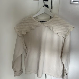 Beige tröja med ribbad krage från Zara - Säljer en beige långärmad tröja från Zara med en bred, ribbad och vågformad krage som ger en unik look. Tröjan har mjukt material och muddar vid ärmsluten. Perfekt för dig som gillar detaljer och vill sticka ut lite extra.