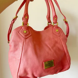 Marc by Marc Jacobs rosa axelväska - Snygg rosa axelväska från Marc by Marc Jacobs i mjukt skinn med guldfärgade metalldetaljer. Väskan har dubbla handtag, justerbar axelrem och en innerficka med dragkedja. Insidan har coolt grafiskt mönster med logotyp. Bredden är ca 36 cm