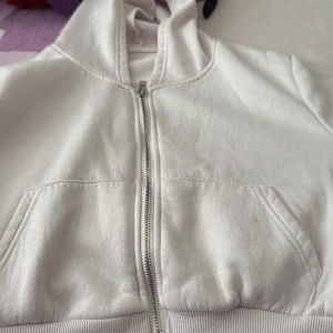 Vit hoodie med dragkedja och huva - Säljer en vit hoodie med huva och praktisk dragkedja framtill. Tröjan har två fickor på magen och ribbade muddar vid nederkant. Perfekt för chill dagar och enkel att matcha med jeans eller mjukisbyxor.